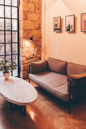 A cozy corner of a living room featuring a mid-century modern style sofa, a terrazzo coffee table with a potted plant, and a tripod floor lamp. The room has a warm ambiance with a combination of a stone wall and a large window with black grid frames. Three framed artworks are hung on the wall above the sofa.