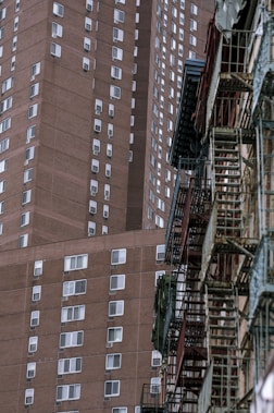 A tall, brown brick residential building with numerous windows, adjacent to an older structure featuring metal fire escapes. The scene captures the urban architecture and density of a city environment.