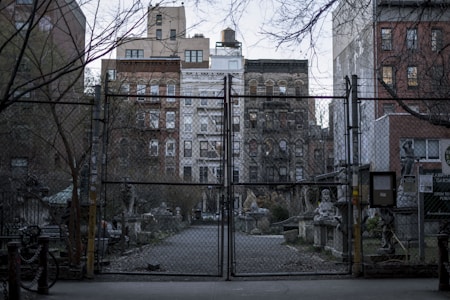 A gated area with a chain-link fence in front of an urban community garden. Several old buildings are visible in the background, surrounded by bare trees and vegetation. Stone sculptures and statues are present near the entrance and along the pathway inside the garden.