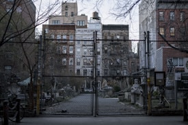 A gated area with a chain-link fence in front of an urban community garden. Several old buildings are visible in the background, surrounded by bare trees and vegetation. Stone sculptures and statues are present near the entrance and along the pathway inside the garden.