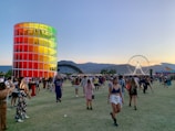 A vibrant outdoor festival scene with people walking and gathering on a vast grassy area during sunset. A tall, cylindrical structure with colorful panels stands prominently on the left. A large Ferris wheel is visible in the background against a backdrop of mountains. Attendees are casually dressed and engaged in various activities.