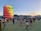A vibrant outdoor festival scene with people walking and gathering on a vast grassy area during sunset. A tall, cylindrical structure with colorful panels stands prominently on the left. A large Ferris wheel is visible in the background against a backdrop of mountains. Attendees are casually dressed and engaged in various activities.