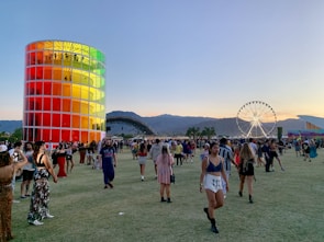 A vibrant outdoor festival scene with people walking and gathering on a vast grassy area during sunset. A tall, cylindrical structure with colorful panels stands prominently on the left. A large Ferris wheel is visible in the background against a backdrop of mountains. Attendees are casually dressed and engaged in various activities.