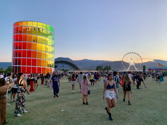 A vibrant outdoor festival scene with people walking and gathering on a vast grassy area during sunset. A tall, cylindrical structure with colorful panels stands prominently on the left. A large Ferris wheel is visible in the background against a backdrop of mountains. Attendees are casually dressed and engaged in various activities.