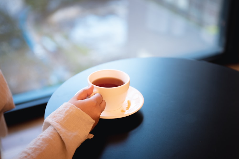 person holding white ceramic teacup on saucer on top of black table