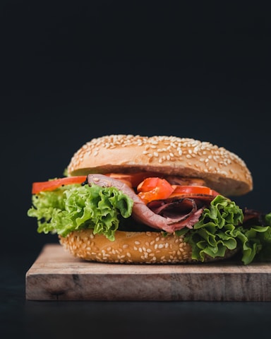 A fresh, gourmet sandwich is placed on a wooden board. The sandwich consists of a sesame seed bun filled with leafy green lettuce, slices of tomato, and layers of roast beef. The vibrant colors of the ingredients stand out against a dark background.