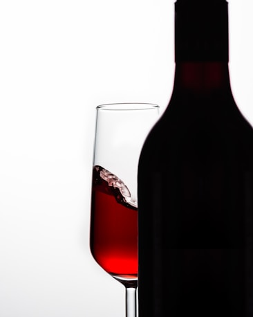 A wine glass partially filled with red wine is positioned behind a silhouetted wine bottle. The glass shows a splash of wine, with a gradient of red tones, set against a light background.