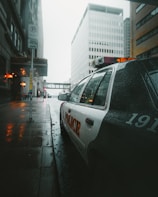 A rainy urban street scene with a police car parked along a wet sidewalk. Tall buildings with modern architecture line the street. Streetlights cast a warm glow, reflecting off the wet pavement. The atmosphere is quiet and the sky is overcast.