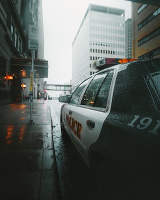A rainy urban street scene with a police car parked along a wet sidewalk. Tall buildings with modern architecture line the street. Streetlights cast a warm glow, reflecting off the wet pavement. The atmosphere is quiet and the sky is overcast.
