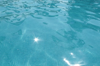 Clear blue water in a swimming pool, with light reflections creating a sparkling effect on the surface. The water appears clean and has a calm texture, with subtle ripples and a grid pattern from the pool tiles visible beneath.