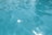 Clear blue water in a swimming pool, with light reflections creating a sparkling effect on the surface. The water appears clean and has a calm texture, with subtle ripples and a grid pattern from the pool tiles visible beneath.