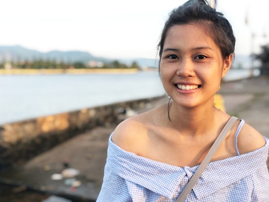 A woman is smiling while standing outdoors near a body of water. She is wearing an off-the-shoulder checkered top and has her hair tied back. The background features a blurred view of water, a stone wall, and distant trees or vegetation.