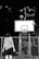 A person is standing in front of a basketball hoop, wearing sports attire. A basketball is captured in mid-air as it approaches the hoop. The scene takes place at night with the city's lights visible in the background.