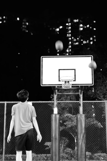 A person is standing in front of a basketball hoop, wearing sports attire. A basketball is captured in mid-air as it approaches the hoop. The scene takes place at night with the city's lights visible in the background.