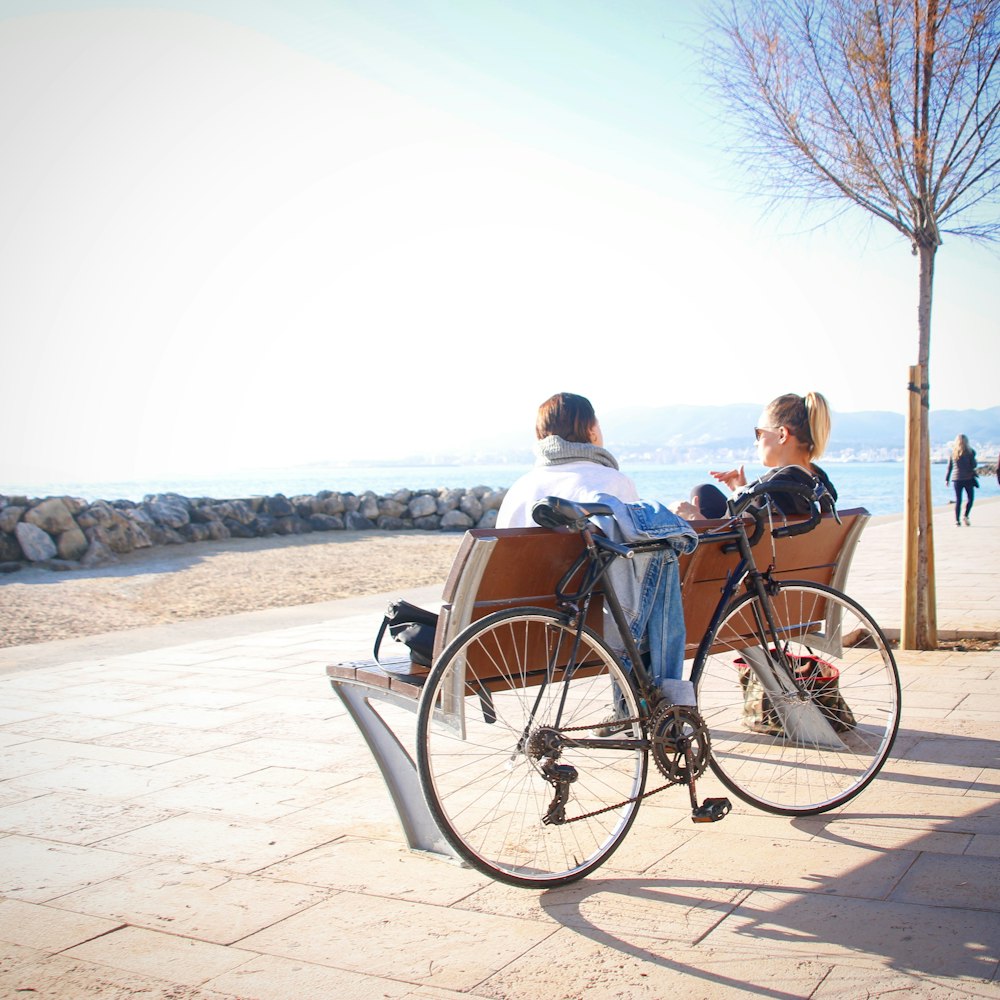 2 person sitting on bench with bike leaning on the back part of bench