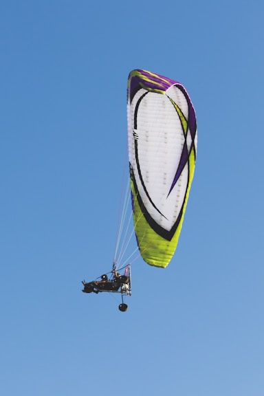A powered paraglider soars against a clear blue sky. The parachute is primarily white with accents of green, yellow, and purple. Two people are seated in the motorized frame suspended below the canopy.