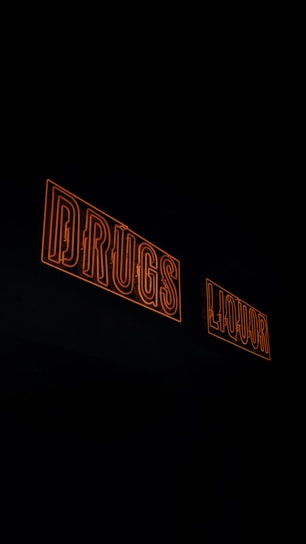 Two neon signs with orange outlines displaying the words 'DRUGS' and 'LIQUOR' against a dark background.