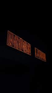 Two neon signs with orange outlines displaying the words 'DRUGS' and 'LIQUOR' against a dark background.