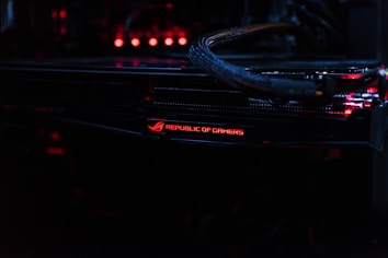 A close-up view of a computer component featuring the logo and lettering 'Republic of Gamers.' The setup is illuminated with red LED lights, and cables are visible connecting the hardware.