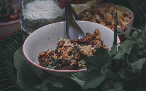 A bowl filled with a savory dish, garnished with herbs and accompanied by metal utensils, is surrounded by green leafy vegetables on a banana leaf. Nearby, there are additional bowls, one containing rice wrapped in transparent plastic and another with crispy-looking elements.