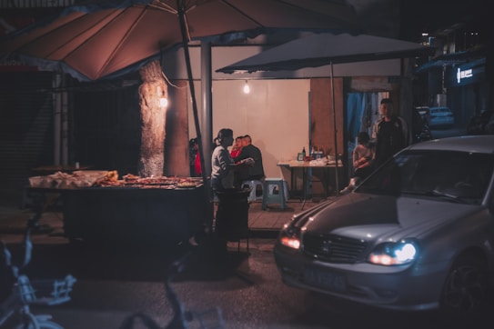 A small outdoor food stall is set up under large umbrellas, illuminated by warm lights. Several individuals are gathered around a table with stools, appearing to be eating or socializing. A car is parked nearby, with its headlights glowing softly. The overall scene suggests an evening market or a street food setup, with a blend of human interaction and urban elements.
