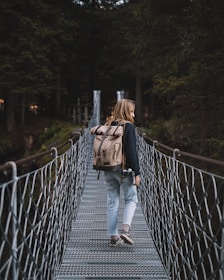 A person with long hair walks across a metal suspension bridge surrounded by tall, dense trees. They are carrying a large beige backpack and holding a camera in one hand. The scene is set in a forest, creating a sense of adventure and exploration.