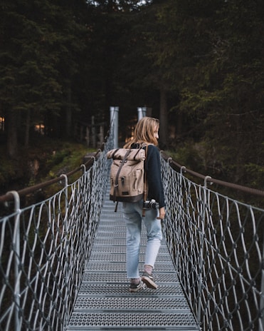 A person with long hair walks across a metal suspension bridge surrounded by tall, dense trees. They are carrying a large beige backpack and holding a camera in one hand. The scene is set in a forest, creating a sense of adventure and exploration.