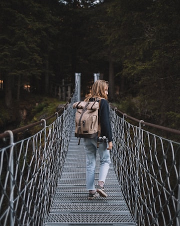 A person with long hair walks across a metal suspension bridge surrounded by tall, dense trees. They are carrying a large beige backpack and holding a camera in one hand. The scene is set in a forest, creating a sense of adventure and exploration.