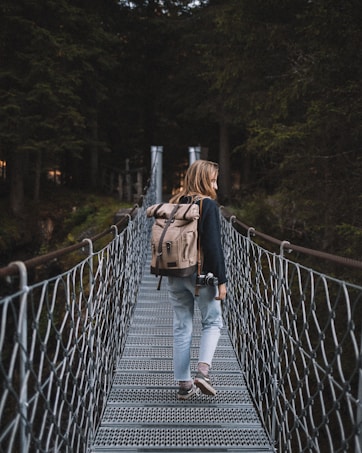 A person with long hair walks across a metal suspension bridge surrounded by tall, dense trees. They are carrying a large beige backpack and holding a camera in one hand. The scene is set in a forest, creating a sense of adventure and exploration.