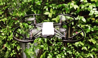 A small drone with propeller guards is hovering in front of a lush green leafy background. The drone features four rotors and appears to be in motion, with blurred propellers indicating flight.