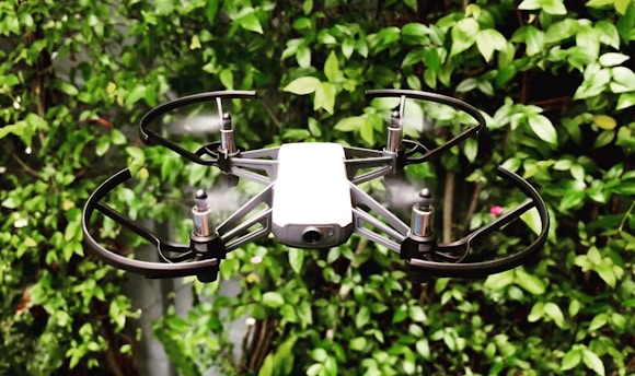 A small drone with propeller guards is hovering in front of a lush green leafy background. The drone features four rotors and appears to be in motion, with blurred propellers indicating flight.