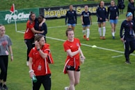 Several women walk across a soccer field wearing sports attire. Some have red shirts, while others are in dark blue warm-ups. They appear to be part of a sports team, likely preparing for or winding down from a soccer match. The background shows a green field with advertisements along the sidelines.