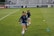 Three athletes are warming up on a grassy field, each wearing navy blue jackets and light blue shorts. They are engaged in a drill involving small orange and blue cones placed on the field. Spectators can be seen sitting in the background in a stadium setting, and advertisements are displayed on the side of the field.