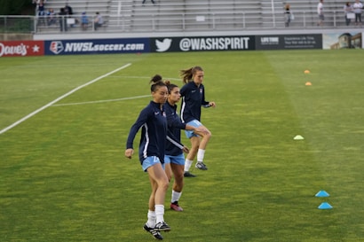 Three athletes are warming up on a grassy field, each wearing navy blue jackets and light blue shorts. They are engaged in a drill involving small orange and blue cones placed on the field. Spectators can be seen sitting in the background in a stadium setting, and advertisements are displayed on the side of the field.