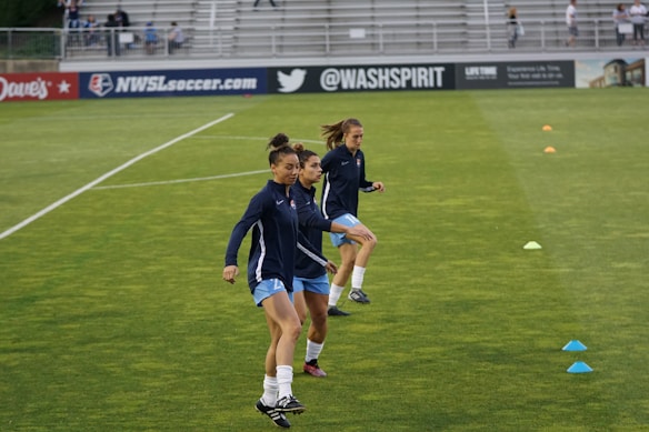 Three athletes are warming up on a grassy field, each wearing navy blue jackets and light blue shorts. They are engaged in a drill involving small orange and blue cones placed on the field. Spectators can be seen sitting in the background in a stadium setting, and advertisements are displayed on the side of the field.