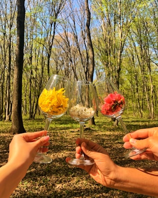 Three hands are holding wine glasses filled with colorful flowers, set against a backdrop of a sunlit forest with tall, leaf-covered trees.