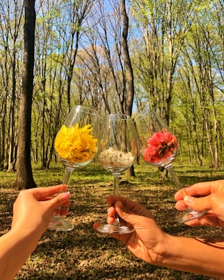 Three hands are holding wine glasses filled with colorful flowers, set against a backdrop of a sunlit forest with tall, leaf-covered trees.