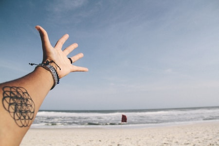 A tattooed and bracelet-adorned hand stretches out towards an expansive beach view with a red flag seen in the distance near the shoreline. The sky is mostly clear with a hint of clouds.