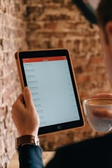 A person holding a tablet displaying a list of items in a minimalistic app interface. The individual is also holding a cup of coffee, suggesting a relaxed setting. The background consists of a textured brick wall, adding a rustic ambiance to the scene.
