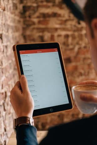 A person holding a tablet displaying a list of items in a minimalistic app interface. The individual is also holding a cup of coffee, suggesting a relaxed setting. The background consists of a textured brick wall, adding a rustic ambiance to the scene.