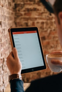 A person holding a tablet displaying a list of items in a minimalistic app interface. The individual is also holding a cup of coffee, suggesting a relaxed setting. The background consists of a textured brick wall, adding a rustic ambiance to the scene.