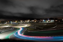 A night view of a racing track with bright, colorful light trails depicting fast-moving vehicles. The surroundings include illuminated buildings and parked cars under a cloudy sky. The track curves prominently in the foreground, creating a dynamic visual effect.