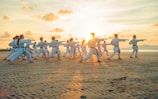 A group of people dressed in martial arts uniforms practice karate on a sandy beach during sunset. They are all in a synchronized stance, performing the same move with extended arms.