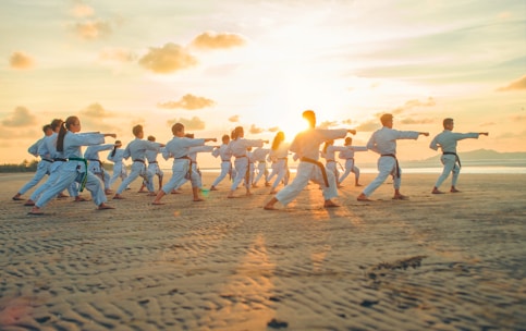 A group of people dressed in martial arts uniforms practice karate on a sandy beach during sunset. They are all in a synchronized stance, performing the same move with extended arms.