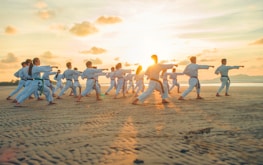 A group of people dressed in martial arts uniforms practice karate on a sandy beach during sunset. They are all in a synchronized stance, performing the same move with extended arms.