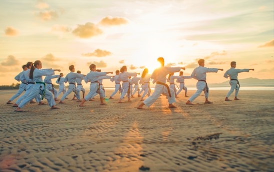 A group of people dressed in martial arts uniforms practice karate on a sandy beach during sunset. They are all in a synchronized stance, performing the same move with extended arms.