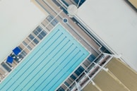 An aerial view of an outdoor swimming pool with multiple lanes in a rectangular shape. The pool area is surrounded by a deck with blue umbrellas and lounge chairs. Adjacent to the pool, a large building with a corrugated roof and several structural elements is visible.