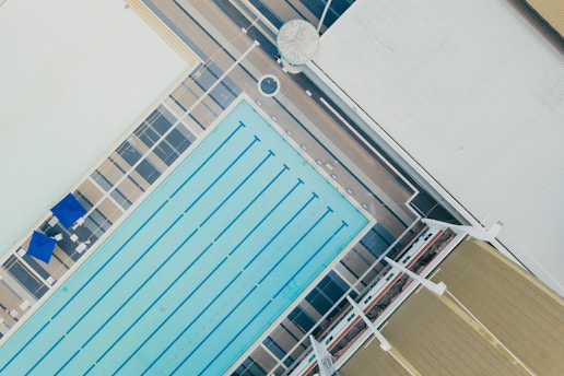 An aerial view of an outdoor swimming pool with multiple lanes in a rectangular shape. The pool area is surrounded by a deck with blue umbrellas and lounge chairs. Adjacent to the pool, a large building with a corrugated roof and several structural elements is visible.
