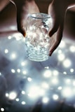 Hands carefully holding a glass jar filled with tiny, glowing fairy lights. The surrounding area is softly illuminated by the warm light emanating from the jar and additional blurred lights create a dreamy atmosphere.
