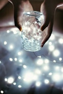 Hands carefully holding a glass jar filled with tiny, glowing fairy lights. The surrounding area is softly illuminated by the warm light emanating from the jar and additional blurred lights create a dreamy atmosphere.